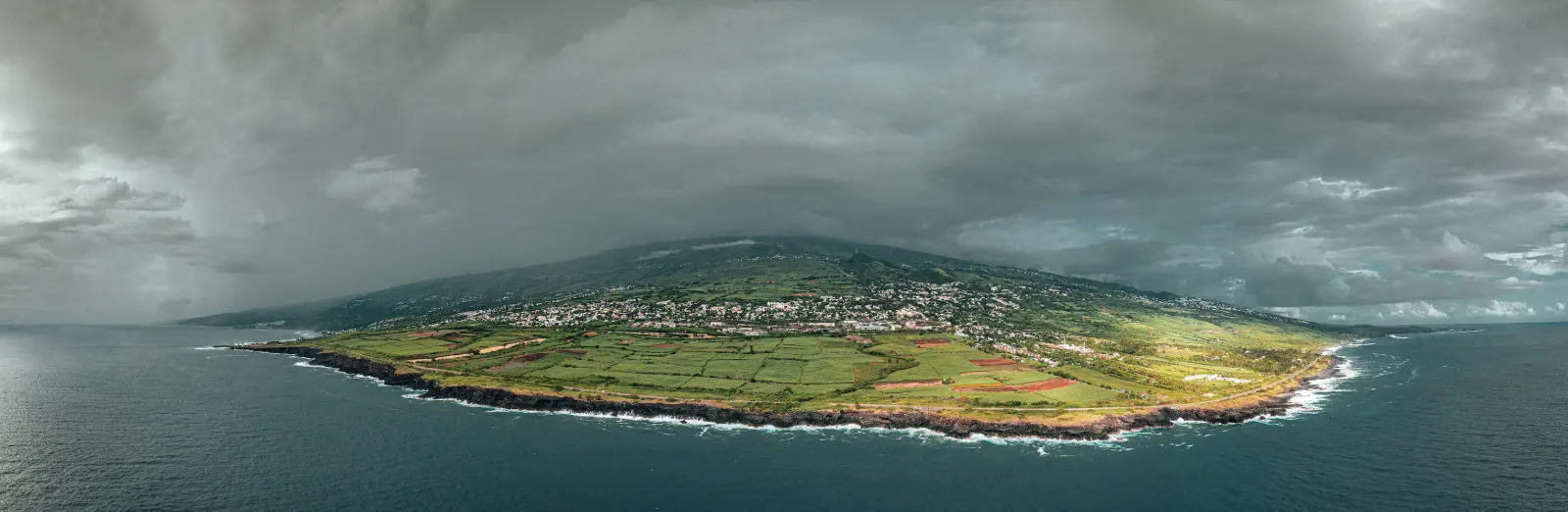 Vue aérienne d'une côte de La Réunion sous un ciel d'orage chargé, champs de canne à sucre, ville et falaises battues par la houle