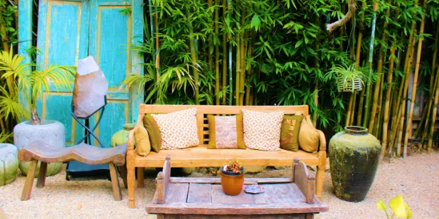 Salon de jardin en bois et coussins devant une haie de bambous et une porte turquoise, coin détente d'un gîte à La Réunion